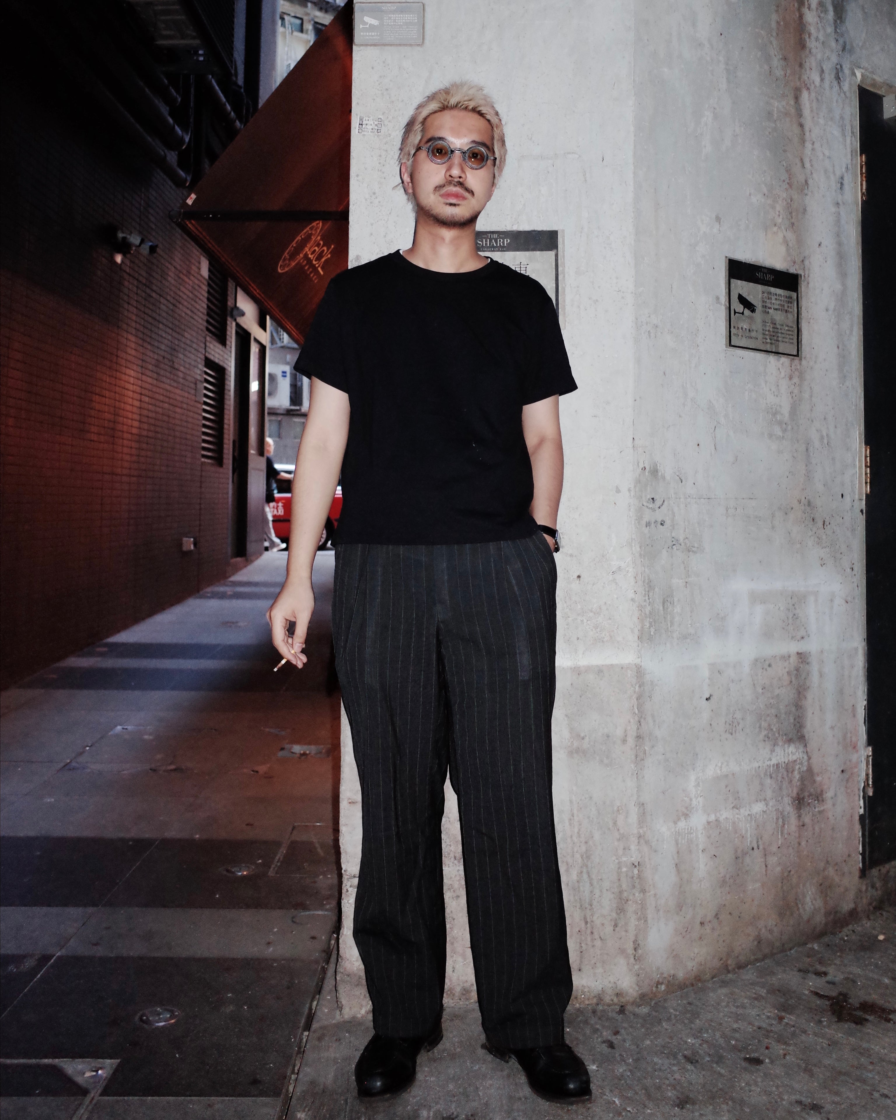 Man wearing a black t-shirt and dark pants standing against a textured wall.