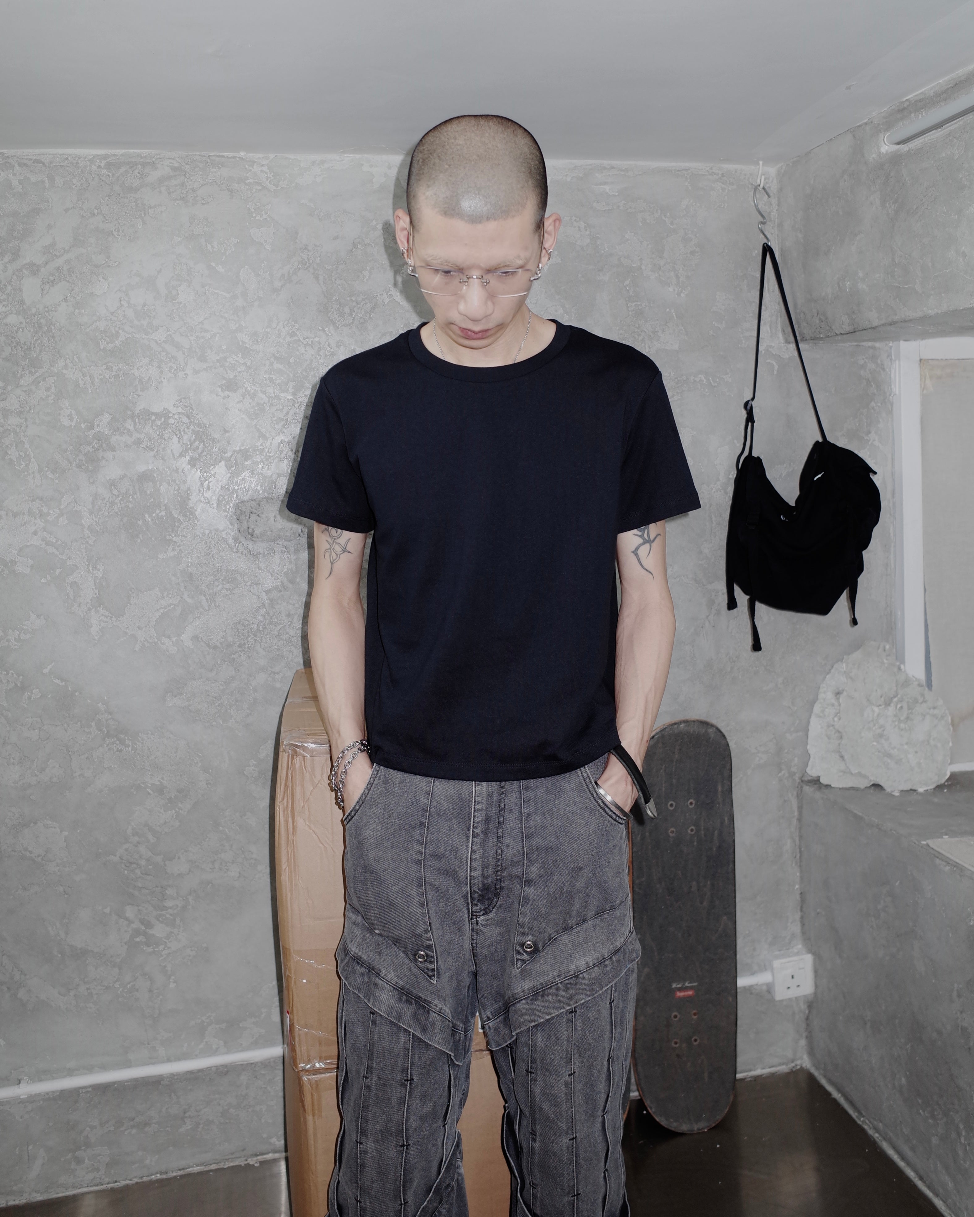 Person wearing a black t-shirt and gray pants standing in a room with concrete walls and a skateboard.