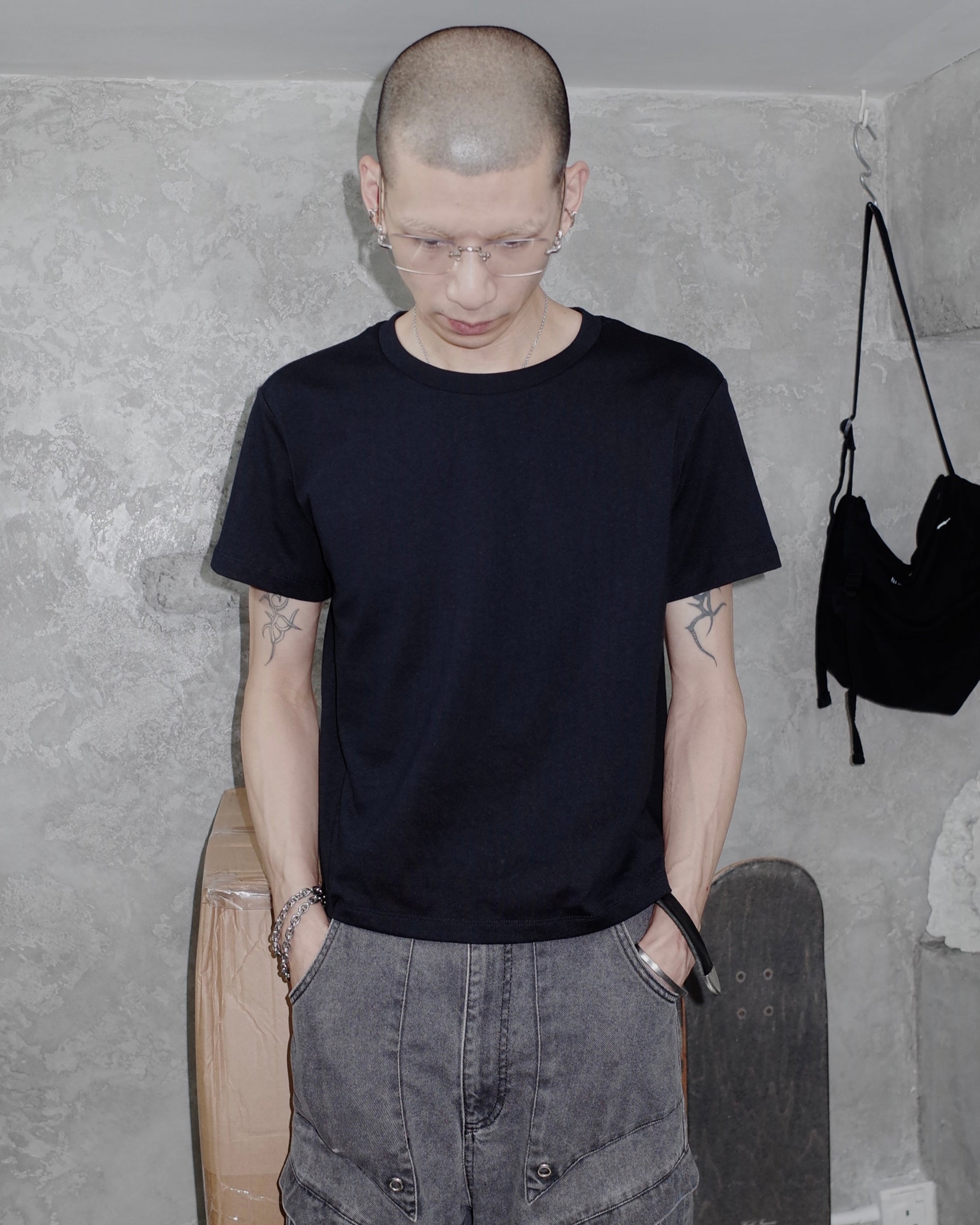 Person wearing a black t-shirt and gray pants standing in a room with concrete walls and a skateboard.