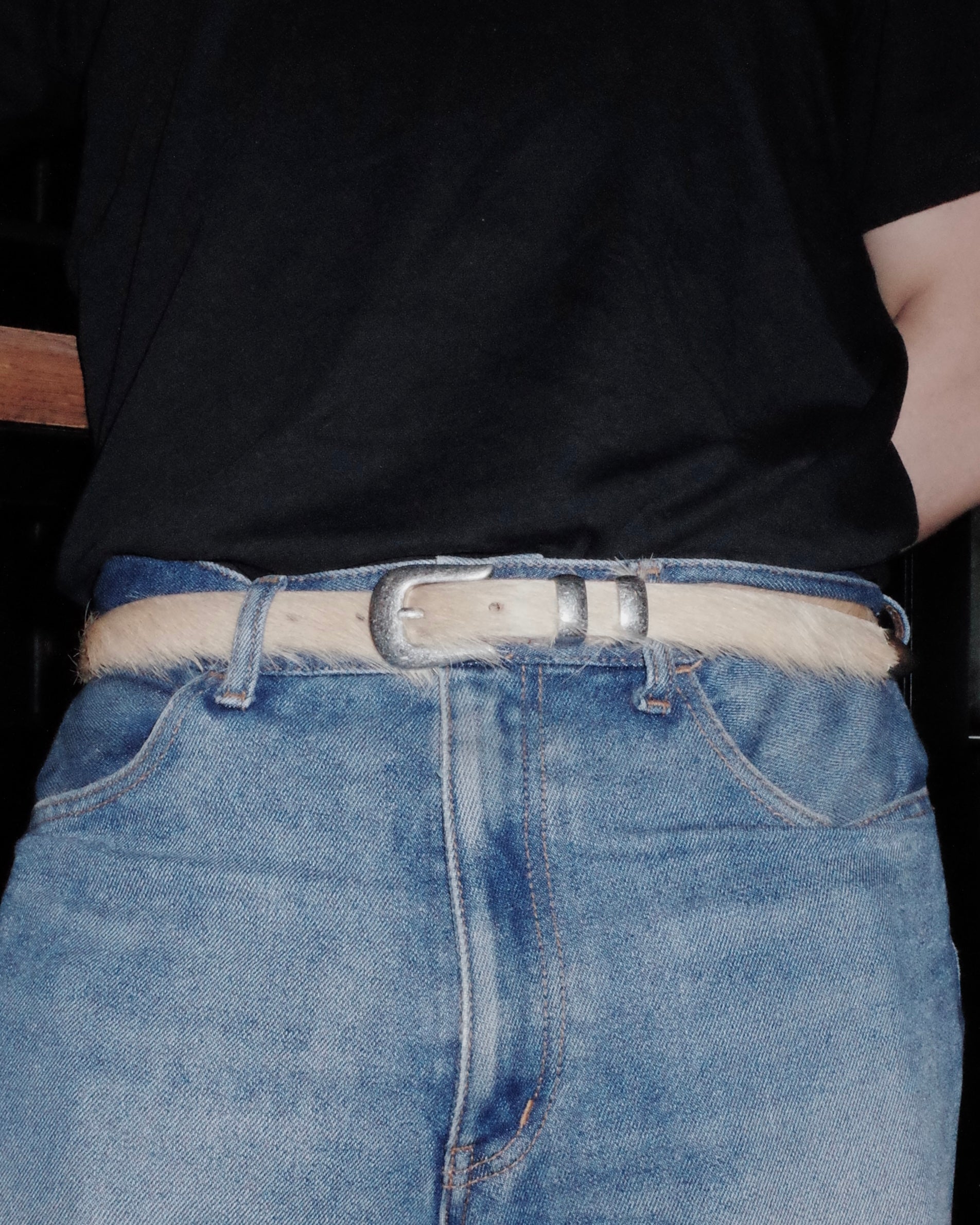Person wearing blue jeans with a beige Western belt and a black shirt.