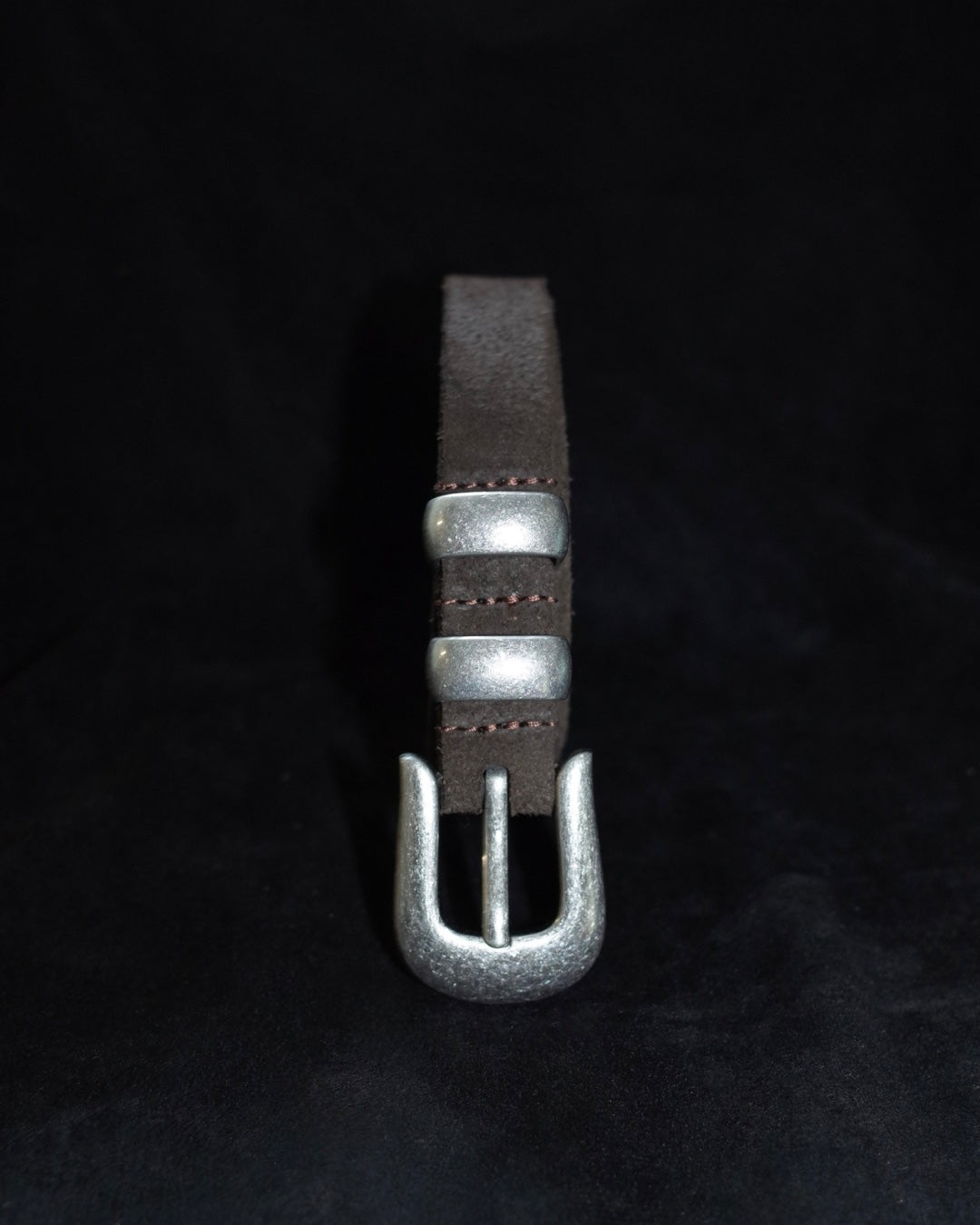 A dark brown leather strap with silver-toned metal hardware, including a textured buckle and two decorative silver rings, displayed vertically against a black background.