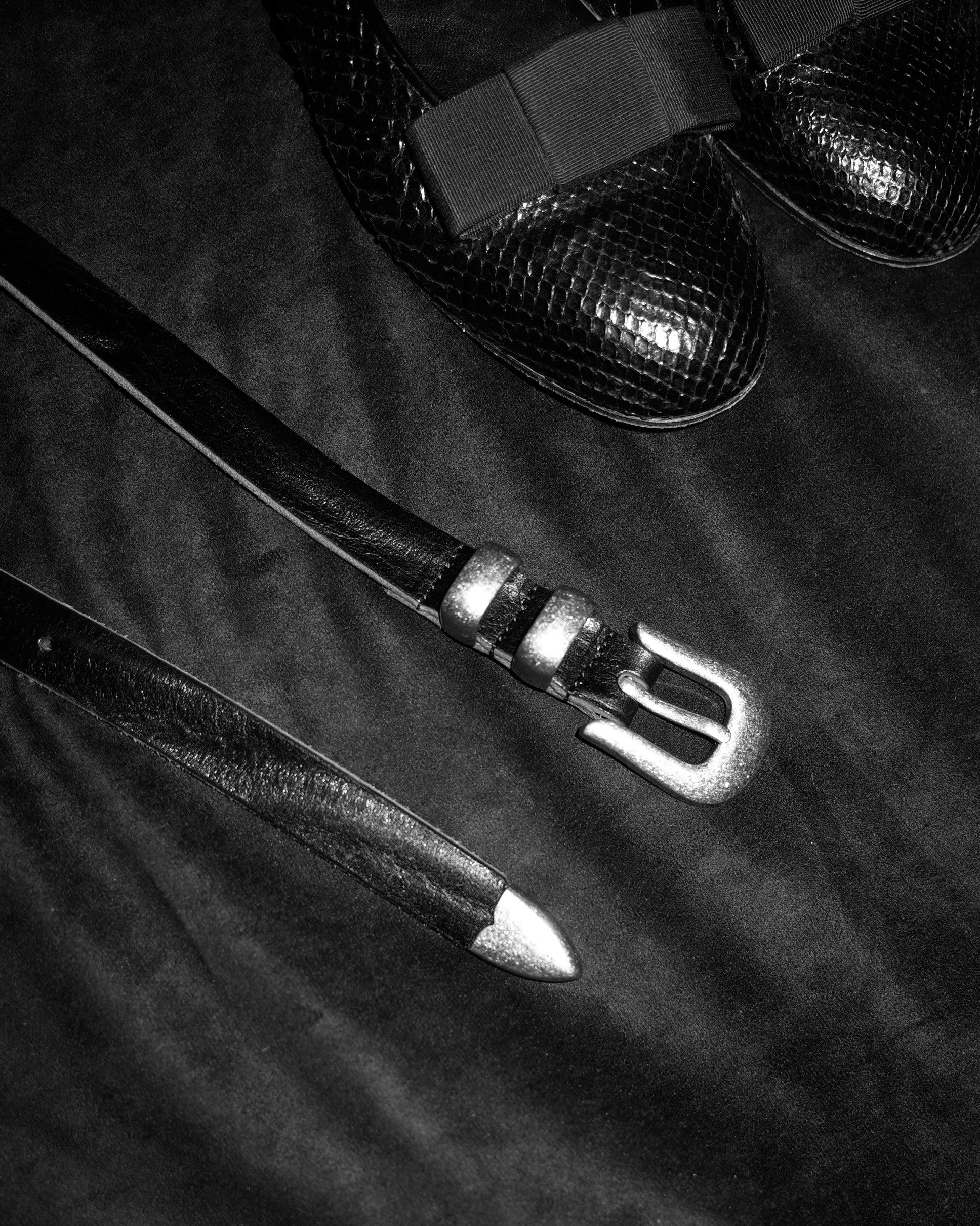 Image of a stylish black leather Western belt with metallic tips and buckles, placed on a dark fabric surface. In the background, there are two black shoes with a snakeskin texture and decorative bows on top.