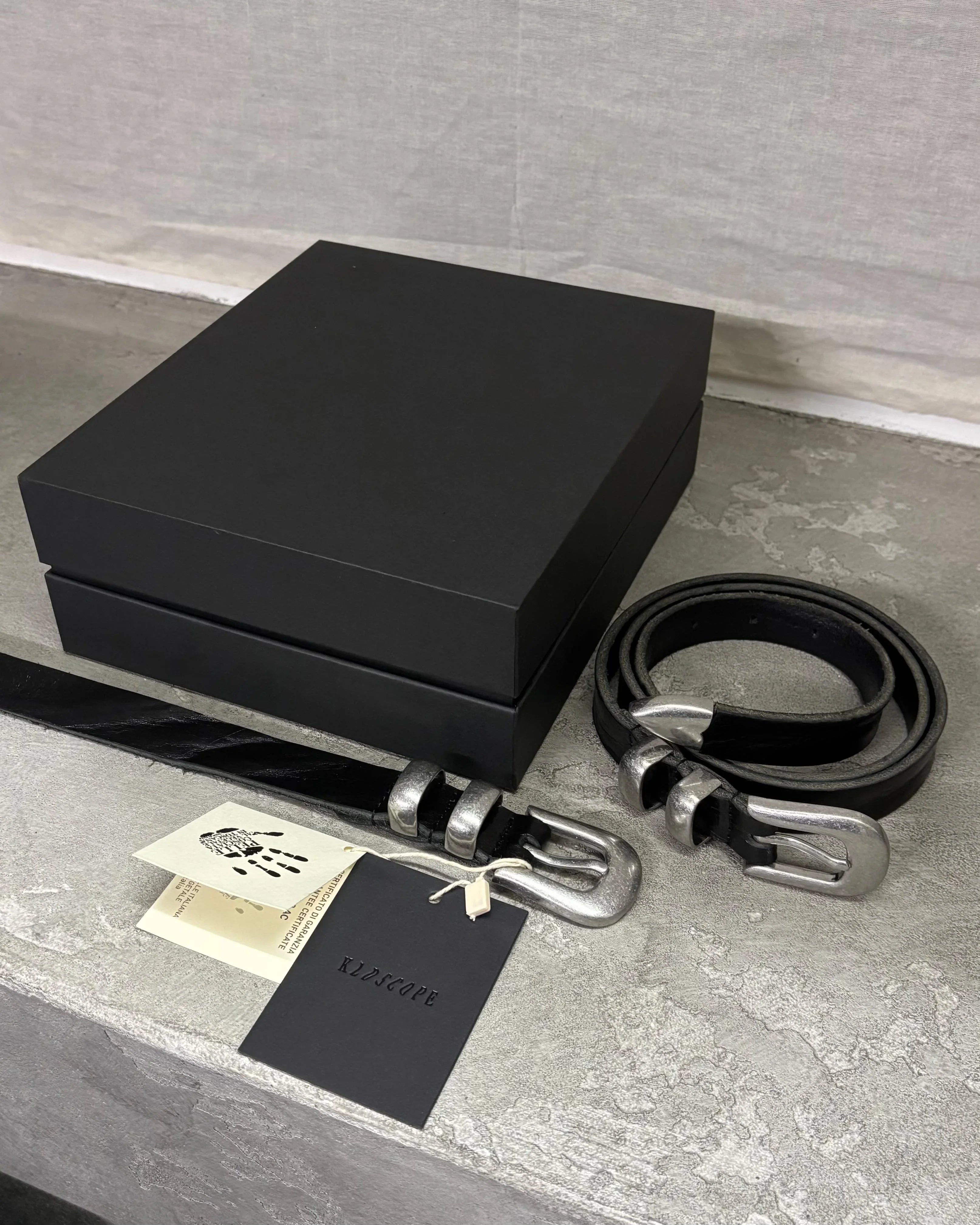 Image of two stylish black leather Western belts with metallic tips and buckles, placed on a stone surface. In the background, there are a box and two hang tags.