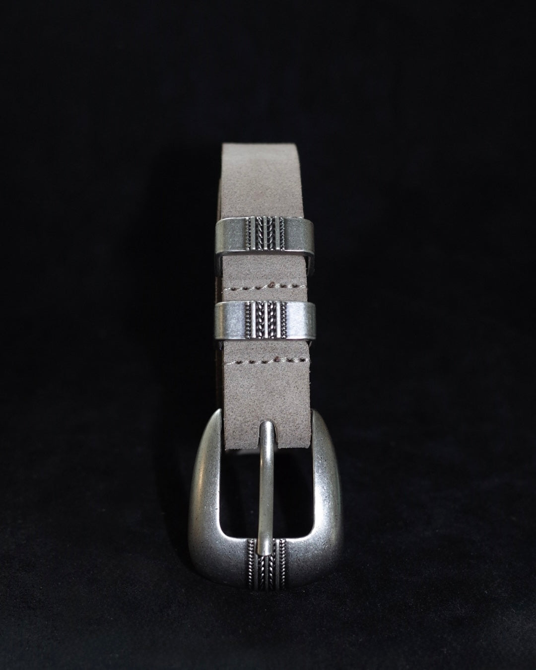 A silver-toned metal belt buckle with engraved decorative details, attached to a light gray leather strap, shown against a solid black background.