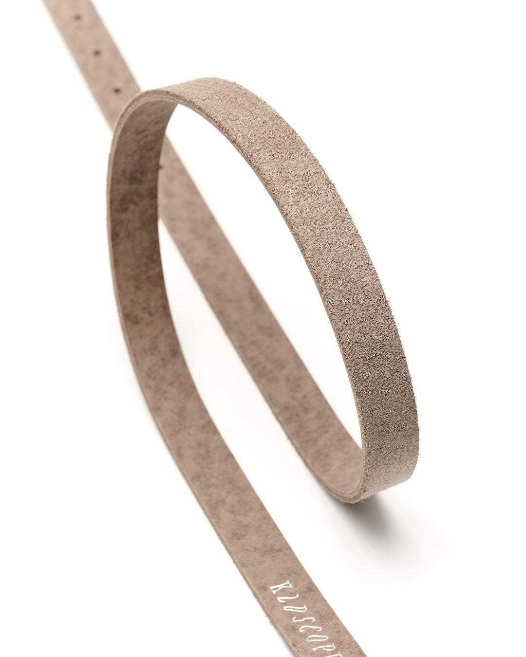 A close-up studio shot of a wide, flat belt made of textured suede or nubuck leather, coiled in an oval shape on a clean white background. The belt is a muted sand-grey colour with a debossed brand name, 'KLDSCOPE ', visible near the bottom.