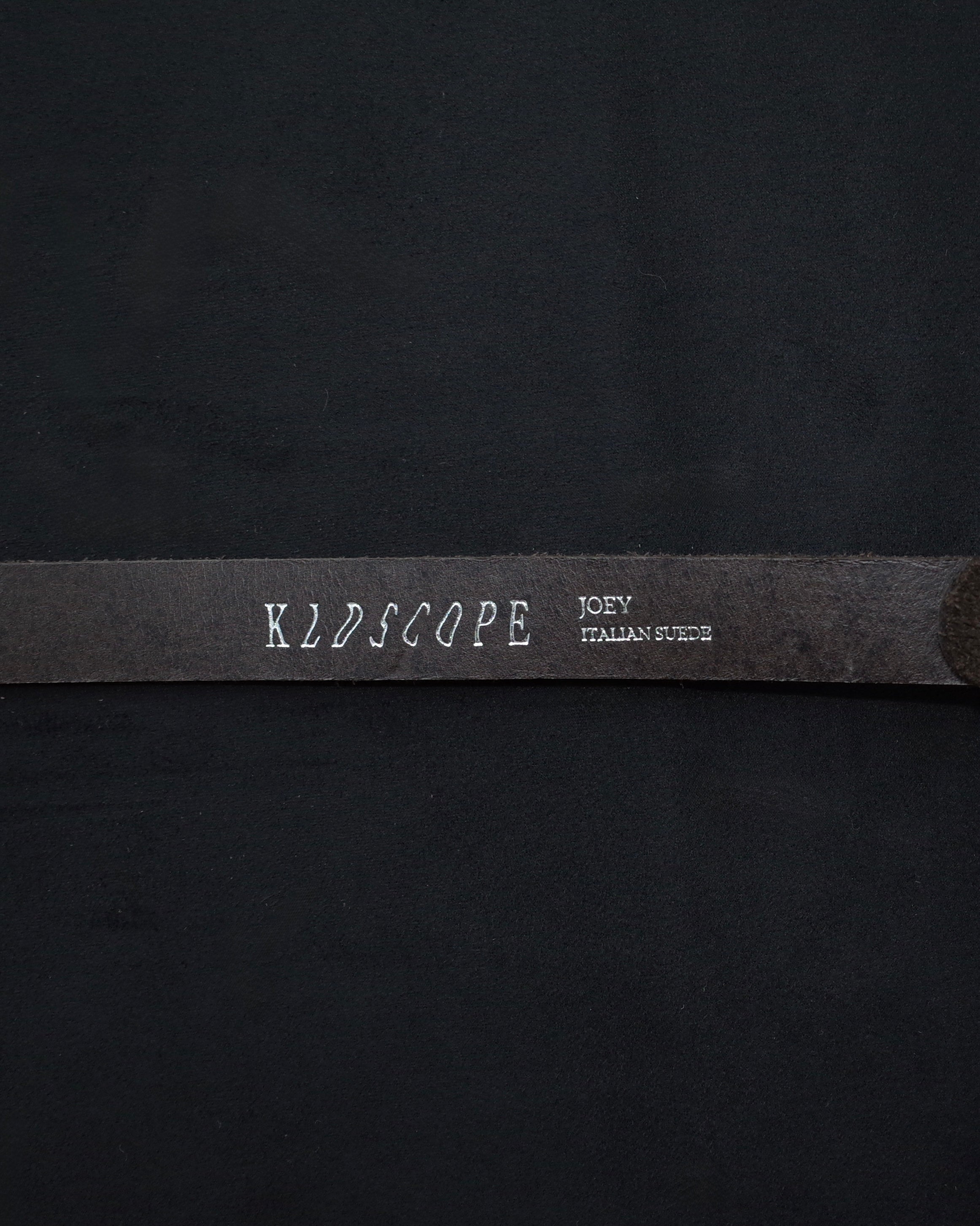 A close-up of a black fabric item, likely an apron, featuring a horizontal black strap with white printed text reading 'KLDSCOPE' and 'JOEY ITALIAN SUEDE'.