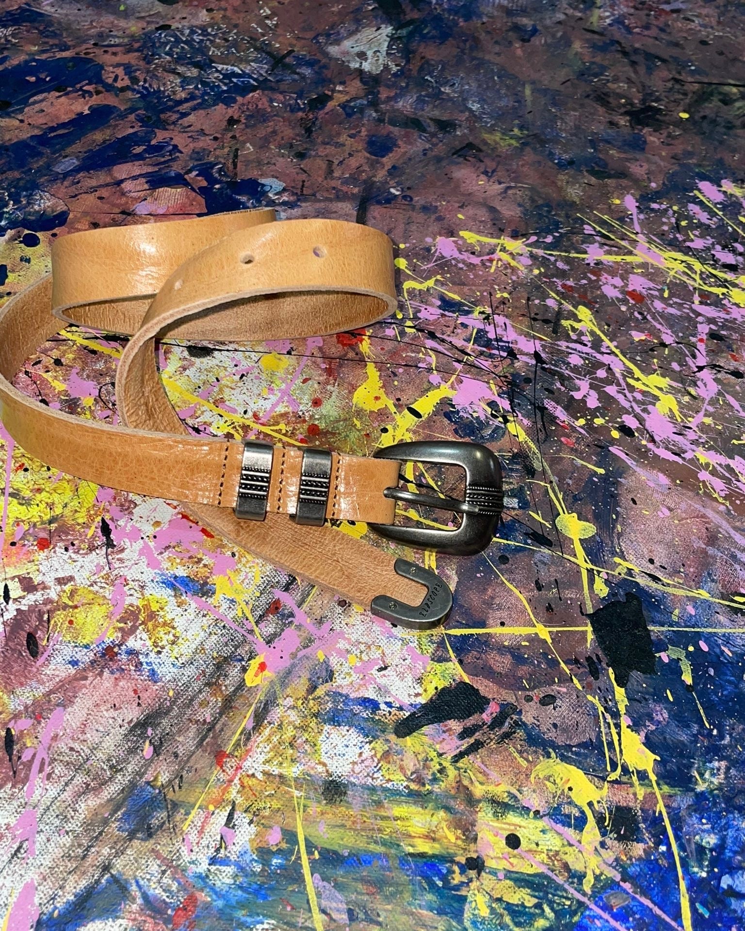 Color image of a tan leather western belt with a metallic buckle and two metal studs, placed on a vibrant abstract background. The background features splashes of paint in colors like blue, yellow, pink, and black, creating a dynamic, textured surface.
