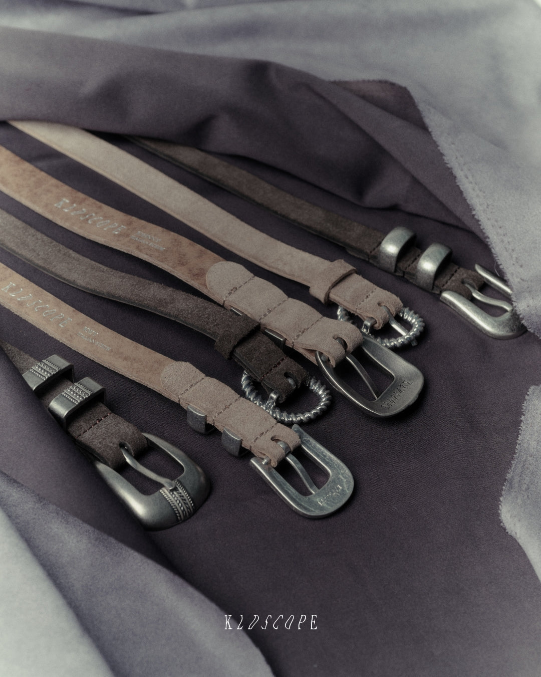 Four leather belts in sand grey and charcoal brown colours with silver-toned metal buckles, arranged diagonally on a dark grey fabric background. The brand name 'KLDSCOPE' is printed on the straps and shown as a watermark at the bottom.